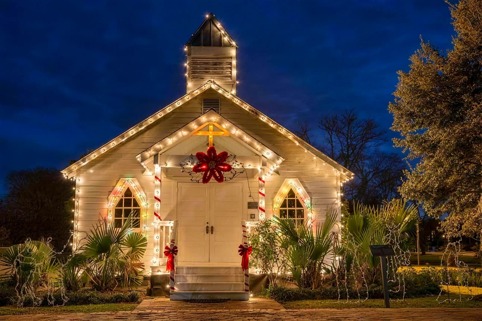 Red brick church with a white steeple outlined in warm white Christmas lights, oak trees on the lawn wrapped in lights