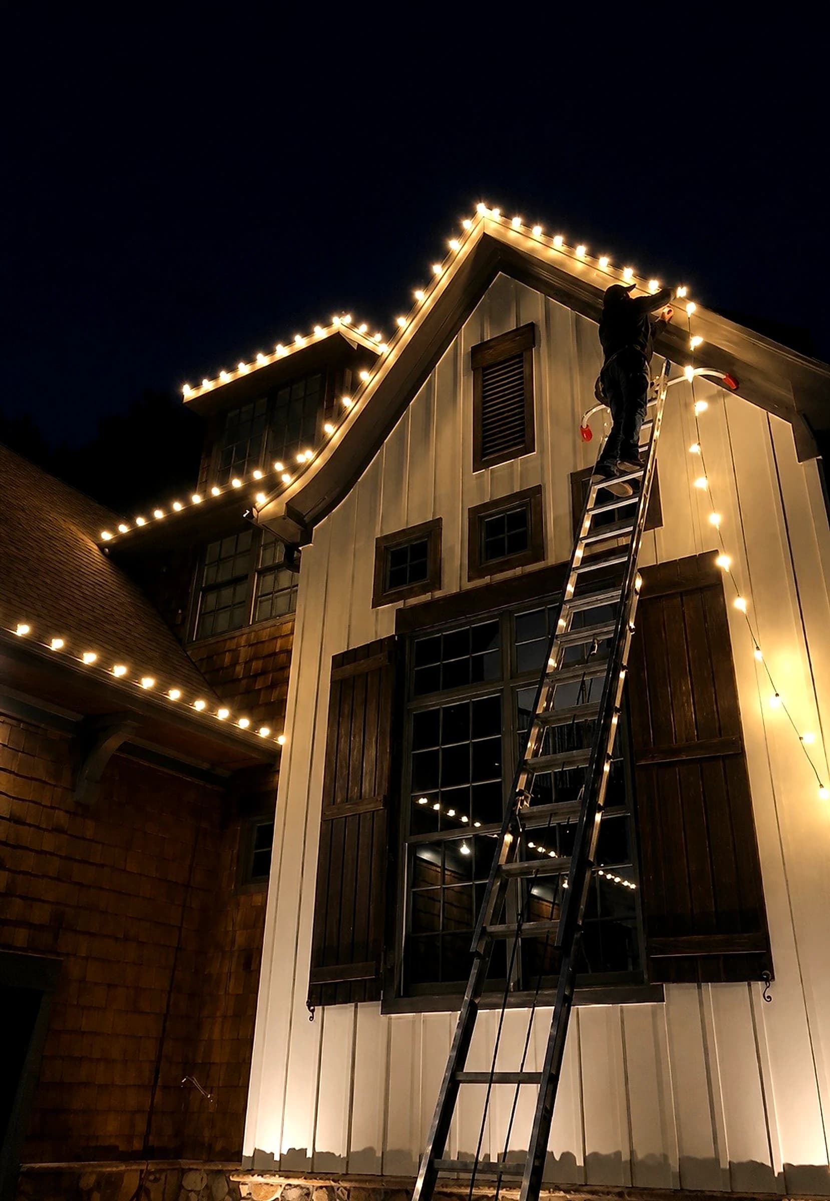 An installer near the top of an extension ladder clipping warm white C9 bulbs along a gable rake two stories up, with the neighboring rooflines already lit