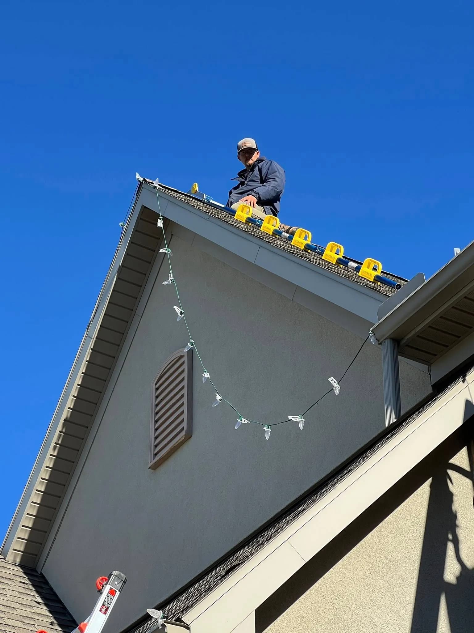 An installer on a steep roof using yellow roof pads and a ladder hook to run a line of C9 bulbs along the gable
