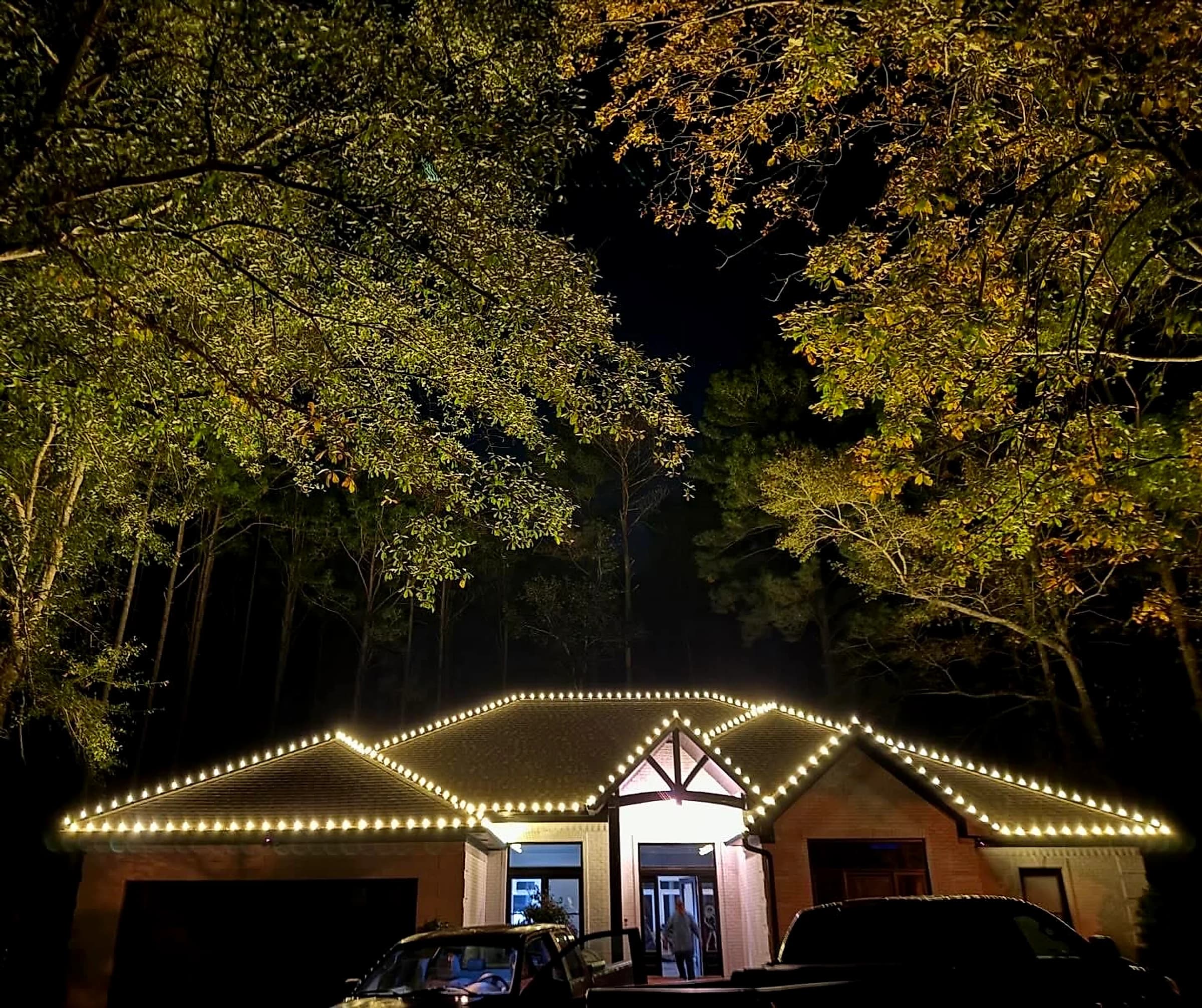 Southeast Lights display in Hattiesburg: a brick home on a wooded lot at night, warm white C9 along every eave, hip and ridge, framed by the tree canopy overhead