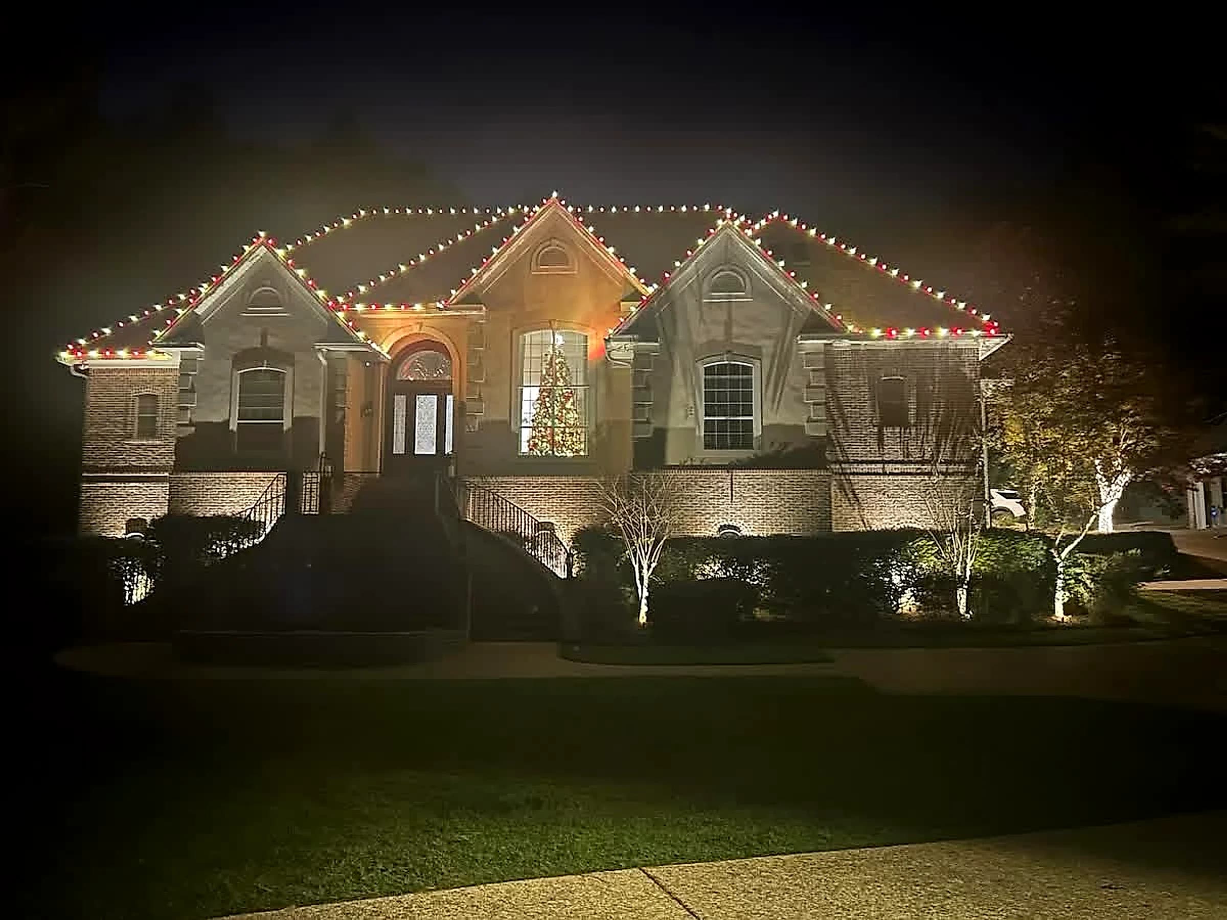 Southeast Lights display in Hattiesburg: a brick home at night with alternating red and warm white C9 bulbs running the full roofline and continuing up every ridge and hip, and a lit Christmas tree visible through the front window