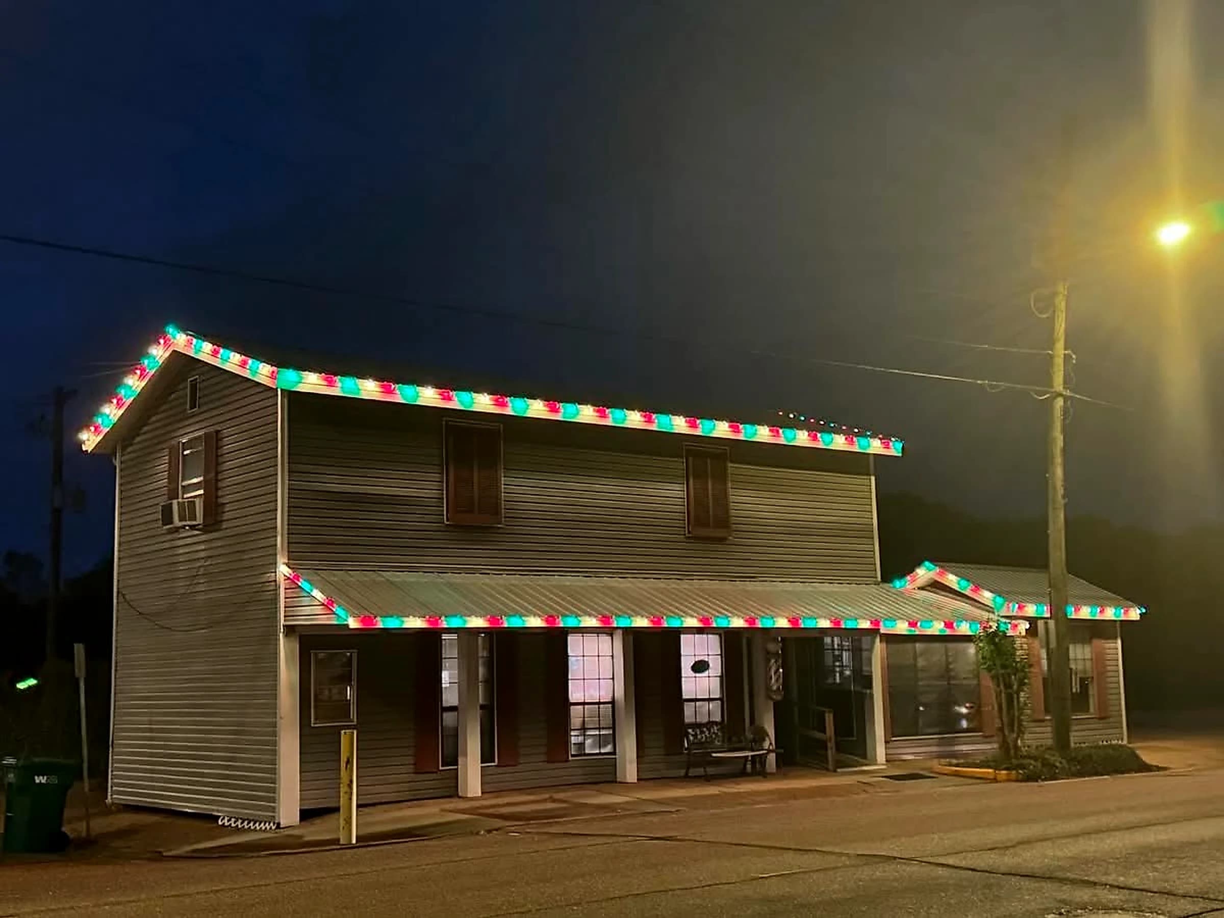 Southeast Lights display in Poplarville: Pearson's Barber Shop at dusk, a two-story Main Street building with red, green and white C9 along the upper gable rake, the full length of the porch awning and the roofline of the attached wing