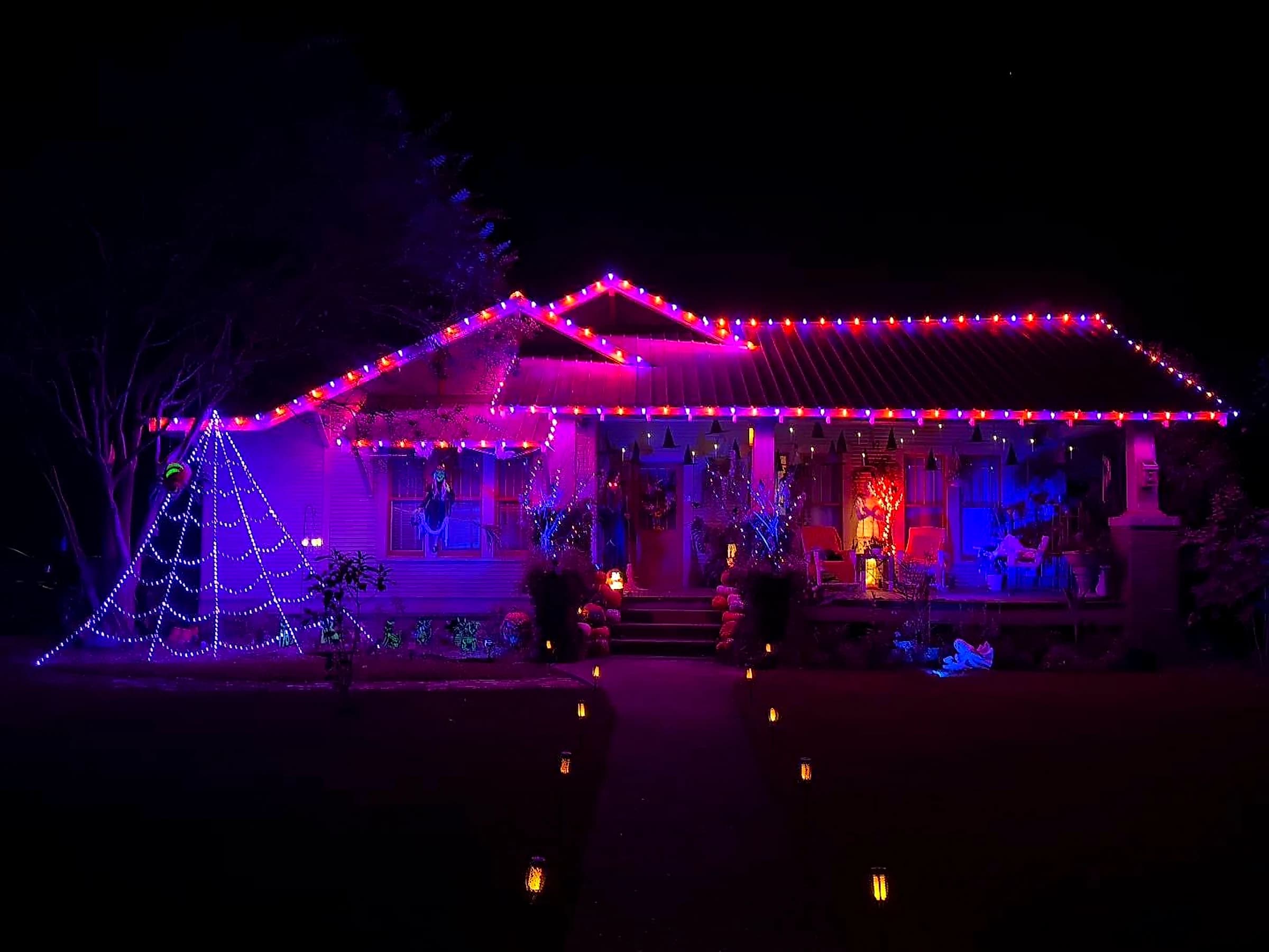 Southeast Lights Halloween display in Poplarville: a bungalow at night with orange and purple C9 bulbs along the roofline, ridges and gable rakes, above a porch decorated for Halloween