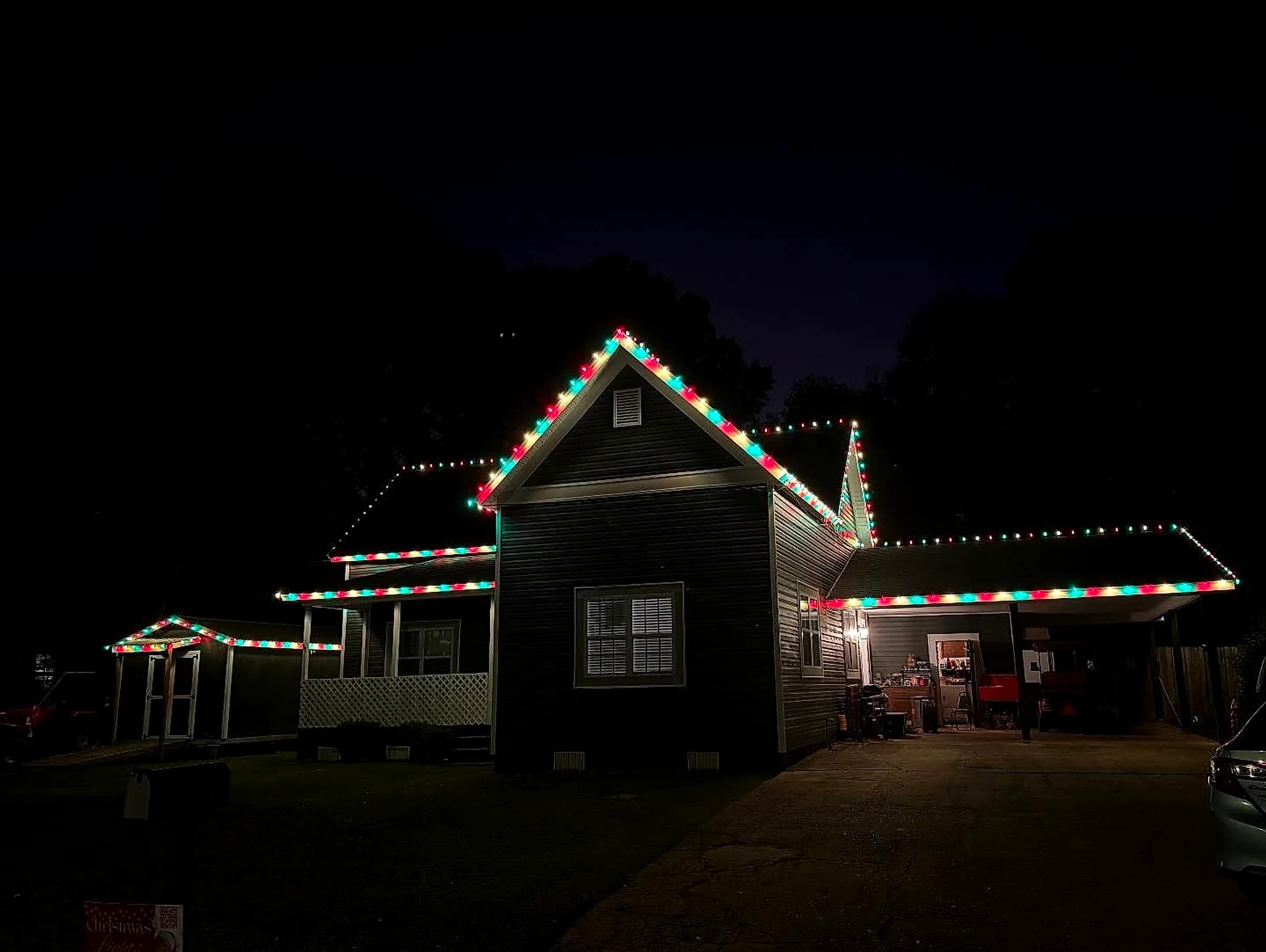 Southeast Lights display in Poplarville: a cottage at night with red, green and white C9 carried around the roofline, up the gable rakes and along the ridges, with the attached carport and a small side building lit in the same pattern