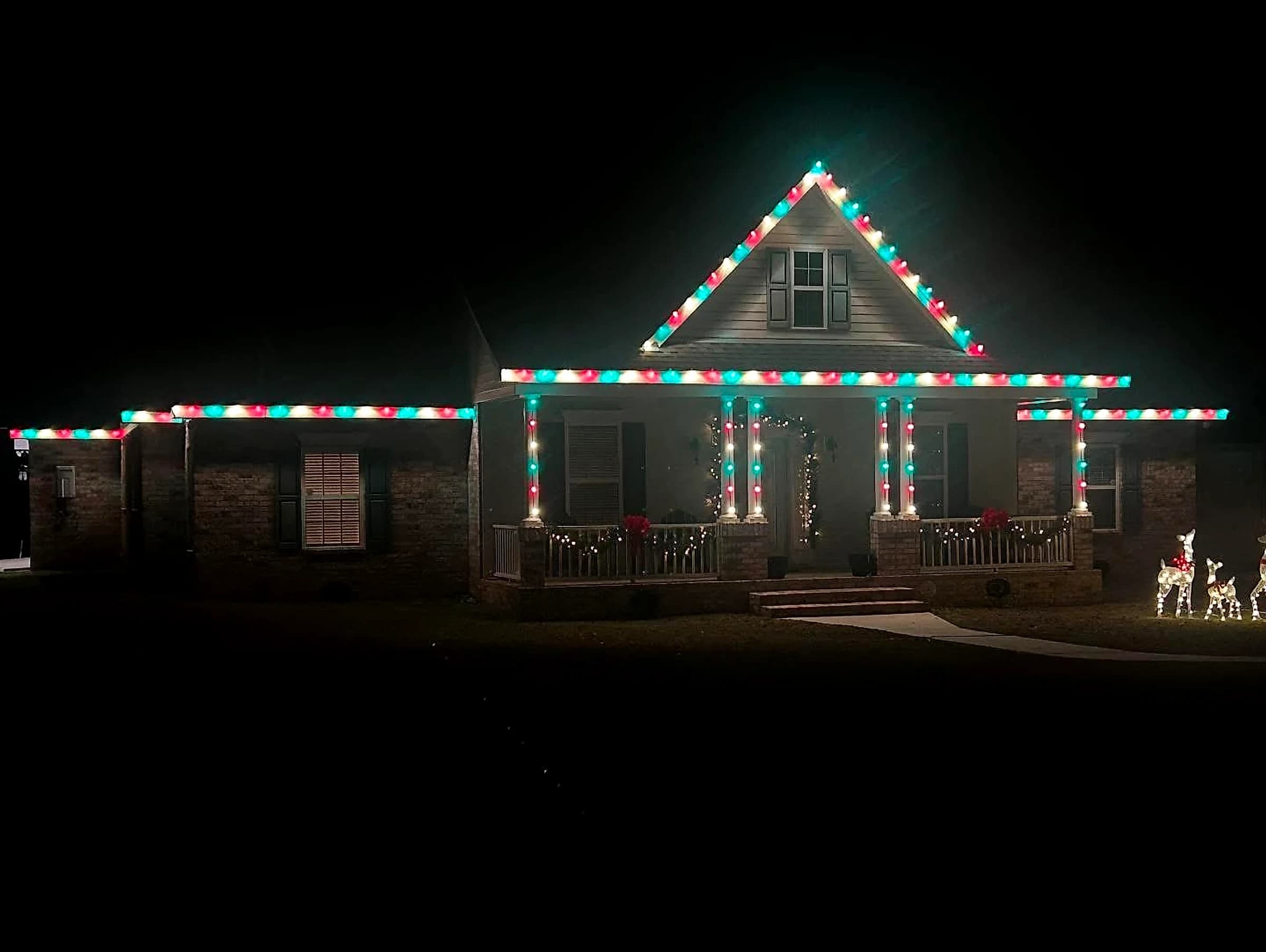 Southeast Lights display in Poplarville: a brick farmhouse at night with a repeating red, green and white C9 pattern across the porch roof, both wing rooflines and the gable rakes, and every porch column wrapped in the same sequence