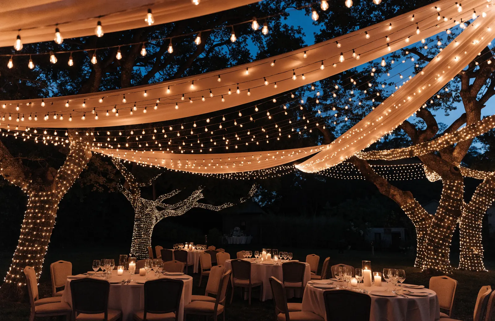 Outdoor wedding reception beneath a canopy of warm white string lights and hanging filament bulbs